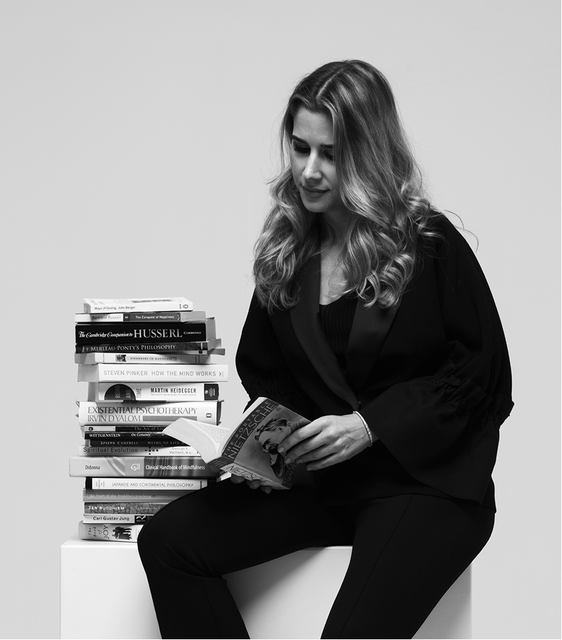 Alt: Dr. Irem Gunay seated beside a stack of psychology and philosophy books, including works by Husserl, Heidegger and Nietzsche.